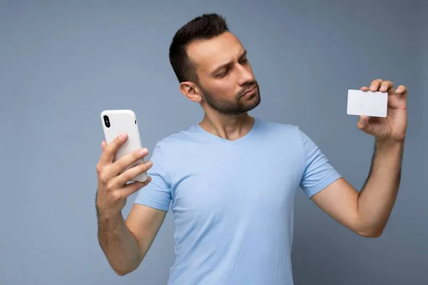 Retrato del hombre guapo concentrado que usa ropa cotidiana aislada en la pared de fondo sosteniendo y usando el teléfono y la tarjeta de crédito haciendo el pago mirando la tarjeta bancaria de plástico —  Fotos de Stock