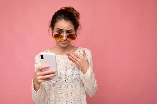 Photo of attractive dissatisfied young woman wearing casual white blouse and colorful sunglasses isolated over pink background wall holding and using phone looking at gadjet display — Stock Photo, Image