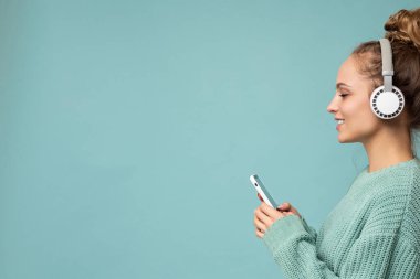 Renkli arka plan duvarından izole edilmiş, beyaz kablosuz kulaklıklar takan, müzik dinleyen ve cep telefonu kullanan şık giyinen güzel bir genç kadının yakın plan fotoğrafı.