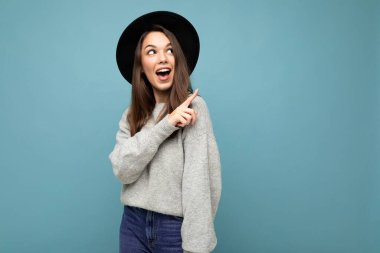 Çekici esmer, mutlu, şaşkın genç bir kadının gri formalı ve mavi arka planda izole edilmiş siyah şapkalı boş bir yeri işaret ettiği fotoğraf.