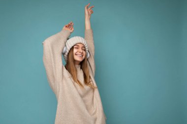 Güzel, mutlu, gülümseyen sarışın bir kadının, bej rengi bir kazak giydiği ve mavi arka planda izole edilmiş bir şapka taktığı kablosuz kulaklık takıp havalı müzik dinlediği bir fotoğraf.