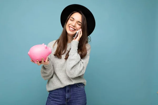Portret zdjęcie szczęśliwego pozytywnego uśmiechu młoda atrakcyjna brunetka nosi swobodny szary sweter i czarny kapelusz odizolowany na niebieskim tle z pustym miejscem, posiada różowy świński grosz bank i mówi — Zdjęcie stockowe