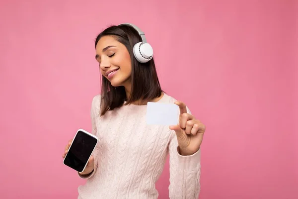 Beautiful young brunette woman wearing pink casual sweater isolated over pink background wall wearing white bluetooth wireless headphones and listening to music and showing mobile phone with empty — Stock Photo, Image