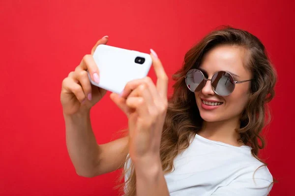 Closeup photo of amazing beautiful young blonde woman holding mobile phone taking selfie photo using smartphone camera wearing sunglasses isolated over colorful wall background looking at device — Stock Photo, Image
