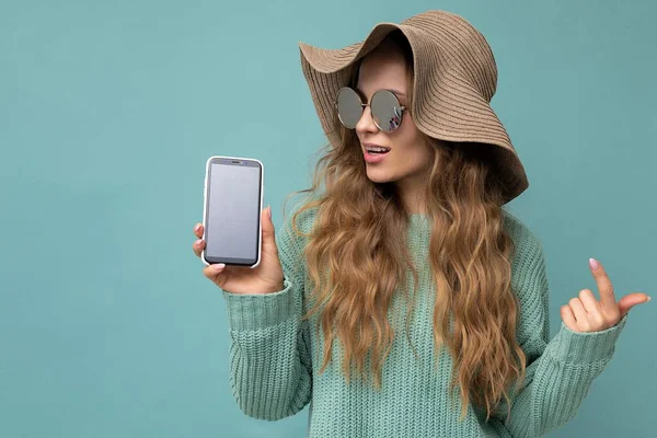 Beautiful happy young blonde woman wearing sunglasses and summer hat isolated on blue background with copy space holding smartphone showing phone in hand with empty screen display looking at camera — Stock Photo, Image