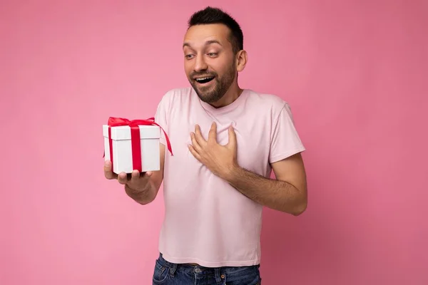 흰색 리본이 달린 흰색 티셔츠를 입고 현재를 보고 있는 핑크 색 티셔츠를 입고 벽에 고립된 수염을 가진 잘생긴 행복 한 젊은 남자의 사진. — 스톡 사진
