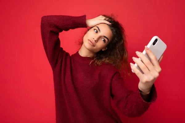Ελκυστική γοητευτική νεαρή χαμογελαστή ευτυχισμένη γυναίκα κρατώντας και χρησιμοποιώντας το κινητό τηλέφωνο λήψη selfie φορώντας κομψά ρούχα απομονώνονται σε φόντο τοίχο — Φωτογραφία Αρχείου