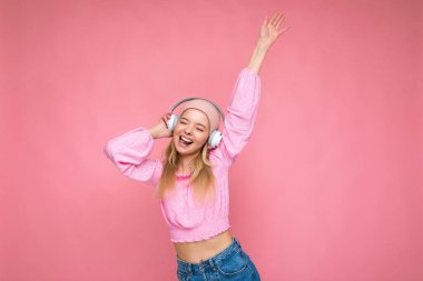 Çekici, pozitif gülümseyen, pembe bluz giyen, pembe şapkalı, arka plan duvarına izole edilmiş, beyaz kablosuz kulaklık takan, müzik dinleyen genç, sarışın bir kadının fotoğrafı.