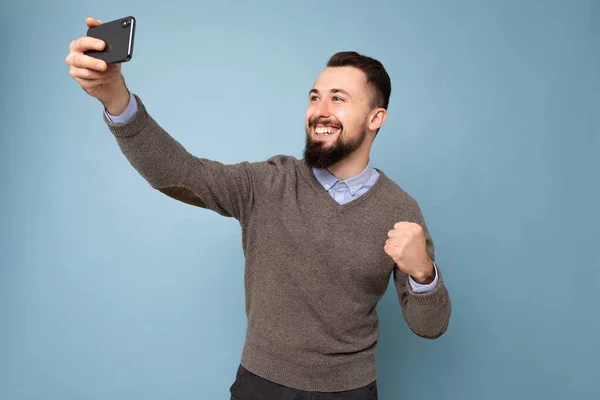 Guapo joven sonriente feliz usando ropa elegante casual de pie aislado sobre la pared de fondo sosteniendo el teléfono inteligente tomando foto selfie mirando la pantalla del teléfono móvil —  Fotos de Stock