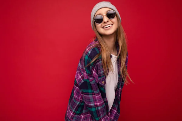 Fascinante mujer rubia oscura joven feliz aislado sobre fondo rojo con camisa casual púrpura sombrero gris y gafas de sol con estilo mirando a la cámara y sonriendo —  Fotos de Stock