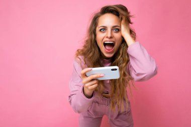 Aşırı mutlu, şaşırmış, pembe kapüşonlu, pembe kapüşonlu, fotokopi çekilen ve cep telefonuyla kameraya bakan güzel, sarışın bir kadının fotoğrafı.