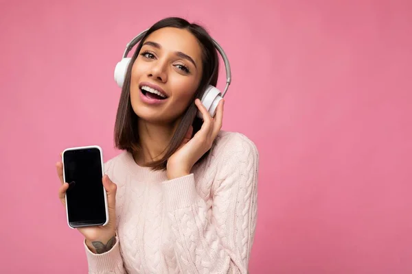 Portret seksowny piękne słodkie młoda brunetka kobieta noszenie różowy sweter izolowane na różowym tle ściany noszenie biały bluetooth bezprzewodowe słuchawki i słuchanie dobrej muzyki i pokazując komórkę — Zdjęcie stockowe