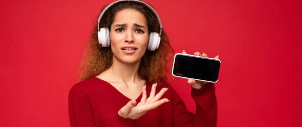 Attrayant insatisfait choqué jeune femme frisée brune portant un pull rouge foncé isolé sur fond rouge portant des écouteurs blancs écoutant de la musique et montrant téléphone moderne avec écran vide pour — Photo