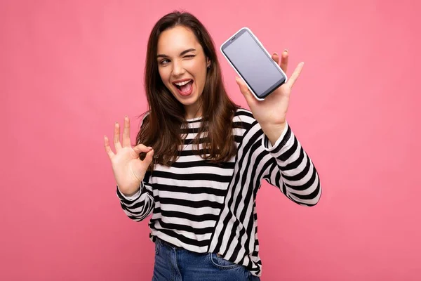 Piscando bela feliz jovem mulher vestindo listrado suéter isolado sobre fundo com espaço de cópia mostrando ok gesto olhando para câmera mostrando tela do telefone móvel — Fotografia de Stock