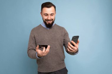 Arkadaki duvarda izole edilmiş günlük giysiler giyen yakışıklı bir adam telefon ve kredi kartı kullanıyor ve akıllı telefon ekranına bakarak ödeme yapıyor.