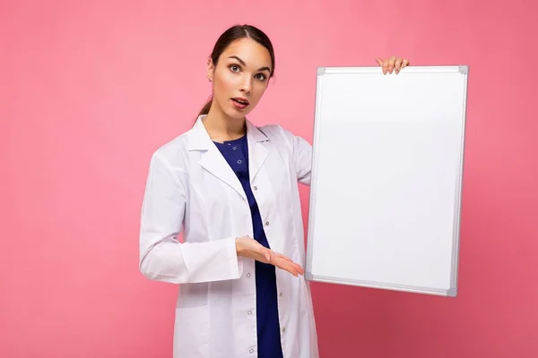 Aufnahme von jungen, hübschen, brünetten Ärztinnen in weißem Mantel mit weißer Magnettafel für Attrappen isoliert auf rosa Hintergrund mit Leerraum — Stockfoto