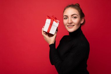 Güzel, mutlu, genç esmer kadın renkli arka plan duvarında izole edilmiş şık günlük kıyafetler giymiş elinde hediye kutusuyla kameraya bakıyor.