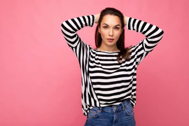 Kendine güveni tam, esmer, samimi duyguları olan, pembe arka planda izole edilmiş, sıradan çizgili kazak giyen güzel bir kadının fotoğrafı.