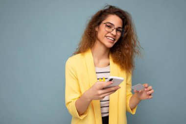 Arka planda izole edilmiş günlük giysiler giyen mutlu genç bir kadın telefon ve kredi kartı ile kredi kartı alışverişini kredi kartıyla yapıyor.