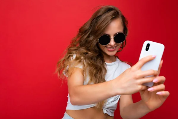 Amazing beautiful smiling young woman holding mobile phone taking selfie photo using smartphone camera wearing sunglasses everyday stylish outfit isolated over colorful wall background looking at — Stock Photo, Image