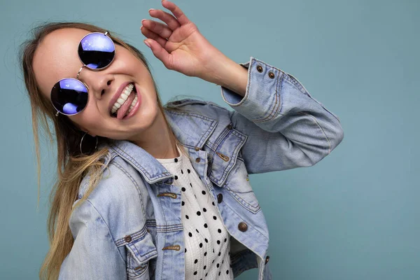 Tiro de mujer rubia divertida divertida bastante positiva con chaqueta de jean azul y gafas de sol elegantes aisladas sobre la pared de fondo azul mirando a un lado y mostrando la lengua — Foto de Stock