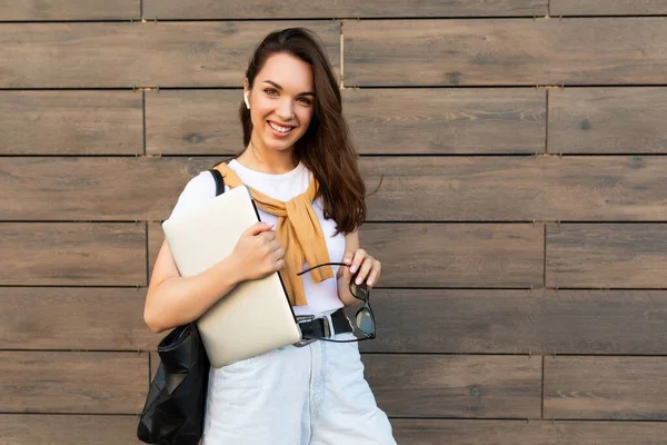 Beautiful smiling charming young brunet woman looking at camera holding computer laptop and sunglasses in white t-shirt and light blue jeans in the street — Stock Photo, Image