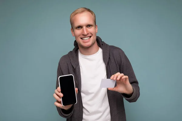 Guapo sonriente hombre feliz con ropa cotidiana aislado en la pared de fondo celebración y el uso de teléfono y tarjeta de crédito haciendo pago mirando a la cámara —  Fotos de Stock