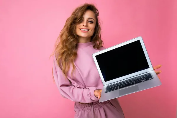 Fotografie de frumos zâmbitor fericit blond cret tânără femeie uita-te la aparat de fotografiat care deține laptop calculator cu ecran monitor gol cu batjocură în sus și copia spațiu purtând hanorac roz izolat peste perete roz — Fotografie, imagine de stoc