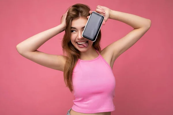 Foto di bella giovane donna bionda positiva che indossa top rosa poising isolato su sfondo rosa con spazio vuoto in mano e mostrando il telefono cellulare con display vuoto per ritaglio cercando di — Foto Stock