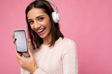 Arka plandaki duvarda izole edilmiş şık giyinen güzel, mutlu, genç bir kadının yakın plan fotoğrafı. Cep telefonunu, beyaz bluetooth giysisiyle boş bir ekran eşliğinde gösteriyor.