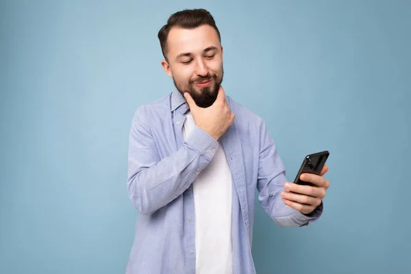 Positive emotional good looking young brunet bearded man wearing casual blue shirt and white t-shirt poising isolated on blue background with empty space holding in hand mobile phone reading sms — Stock Photo, Image