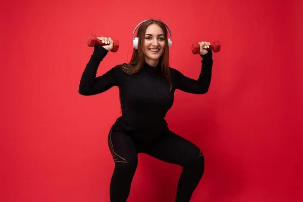 Photo of beautiful happy smiling young brunet woman wearing black sport clothes isolated on red background wall squating wearing white bluetooth headsets listening to music looking at camera — Stock Photo, Image