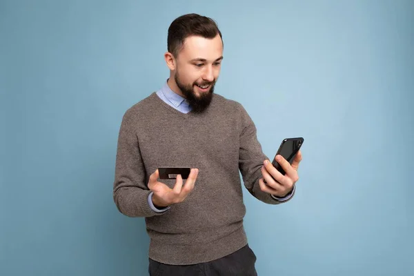 Hombre guapo que usa ropa cotidiana aislada en la pared de fondo sosteniendo y usando el teléfono y la tarjeta de crédito haciendo el pago mirando la pantalla del teléfono inteligente —  Fotos de Stock