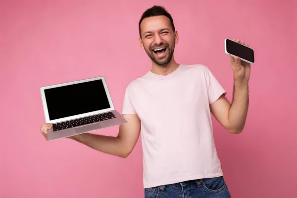 컴퓨터와 휴대 전화를 들고 카메라를 보고 있는 손에 익은 웃음을 보이는 사람 이 고립된 핑크 색 배경 위에 티셔츠를 입고 있다 — 스톡 사진
