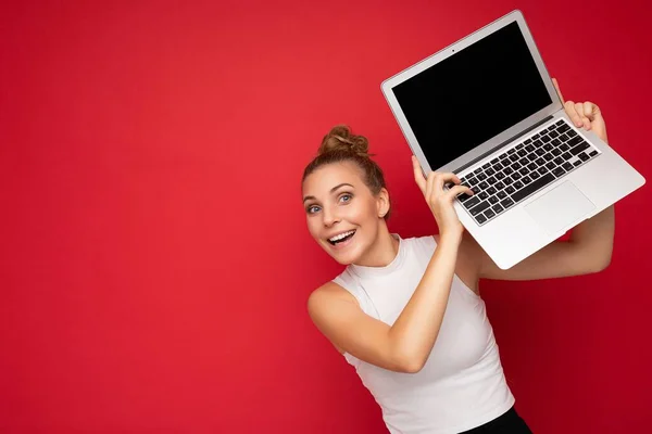 Όμορφη συναρπαστική ευτυχισμένη ξανθιά νεαρή γυναίκα με ανοιχτό στόμα με συγκεντρωμένα μαλλιά κοιτάζοντας κάμερα κρατώντας φορητό υπολογιστή φορώντας λευκό t-shirt απομονώνονται πάνω από κόκκινο φόντο τοίχο — Φωτογραφία Αρχείου