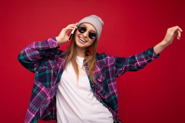 Yakından çekilmiş güzel, mutlu, pozitif sarışın bir kadının hipster mor tişört giydiği, gri tişört ve güneş gözlüğü taktığı fotoğraf. Kırmızı arka planda izole edilmiş, el ele tutuşup,