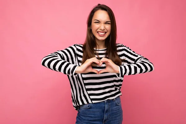 Joven positiva adorable feliz amiling atractiva mujer morena con emociones sinceras usando suéter de rayas casuales aislados sobre fondo rosa con espacio de copia y mostrando el gesto del corazón. Amor. —  Fotos de Stock