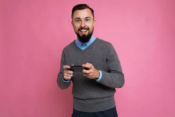 회색 스웨터를 입고 파란 셔츠를 입고 분홍색 배경에 고립되어 빈 공간을 손에 쥐고 휴대 전화를 사용하는 행복해 보이는 잘생긴 수염을 가진 젊은 남자가 웃고 있다. — 스톡 사진
