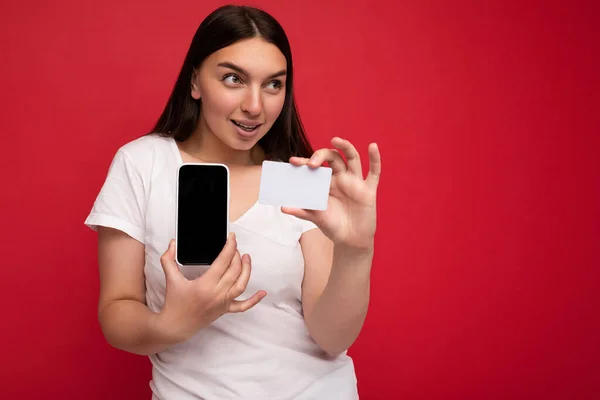 Фото красивой молодой брюнетки в белой футболке на красном фоне с кредиткой и мобильным телефоном с пустым дисплеем для вырезки, смотрящей в сторону — стоковое фото