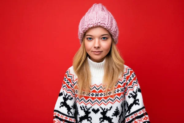 Foto de hermosa calma joven rubia seria aislada sobre la pared de fondo rojo con suéter de invierno y sombrero rosa mirando a la cámara — Foto de Stock