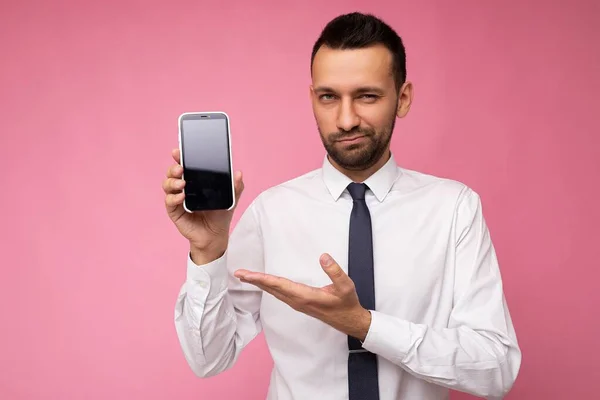 Foto de un hombre guapo seguro de sí mismo con camisa blanca casual y corbata aislada sobre fondo rosa con espacio vacío sosteniendo la mano y mostrando el teléfono móvil con pantalla vacía para maqueta —  Fotos de Stock