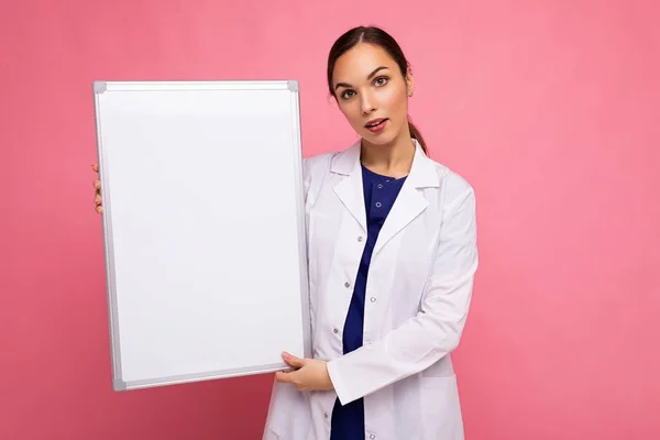 Porträt einer attraktiven hübschen jungen positiven brünetten Frau in medizinischem weißen Mantel mit weißer Magnettafel für Attrappe isoliert über rosa Hintergrund mit Kopierraum — Stockfoto