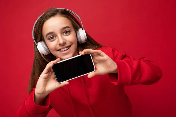 빨간 후드를 입고 빨간 배경에서 고립된 아름다운 갈색 머리 소녀의 클로즈업 사진, 듣고 있는 흰색 무선 헤드셋을 착용 한 스마트폰을 보여 주고 있다. — 스톡 사진