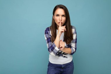 Samimi duyguları olan, mavi arka planda izole ve sessiz bir jest yapan, modaya uygun bir çek tişörtü giyen genç, sinirli, güzel bir esmer kadın. Sessiz ol.