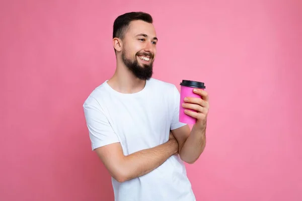 하얀 티셔츠를 입고 핑크 색 배낭 벽에 고립된 채 종이 커피 컵을 들고 옆을 바라보고 있는 건강 한 젊은 갈색 갈색 머리의 남자 — 스톡 사진