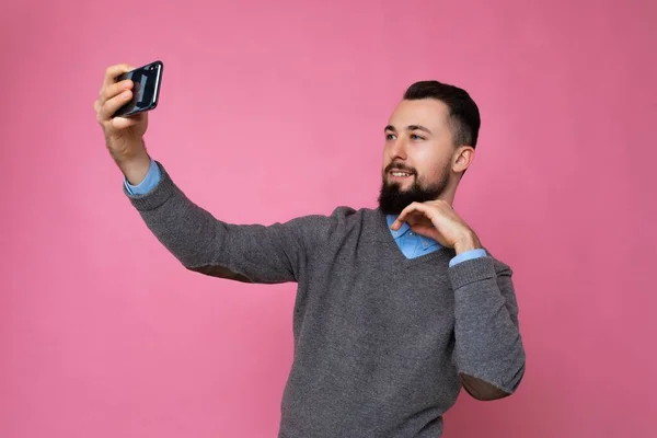 Bonito jovem vestindo roupas casuais e elegantes em pé isolado sobre a parede de fundo segurando smartphone tirar foto selfie olhando para a tela do telefone móvel — Fotografia de Stock