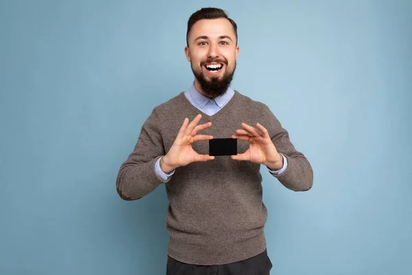 Bonito feliz sorrindo morena barbuda homem pessoa vestindo camisola cinza e camisa azul isolado na parede de fundo segurando cartão de crédito olhando para a câmera — Fotografia de Stock