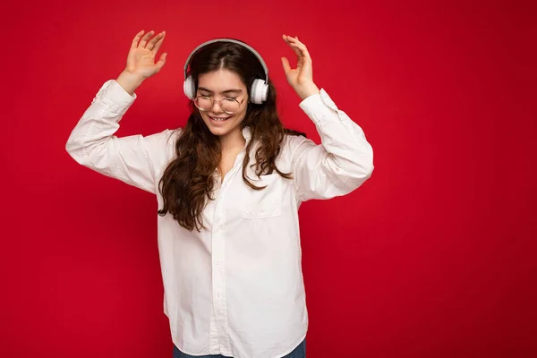 Attraktive, glücklich lächelnde junge brünette Frau mit weißem Hemd und optischer Brille auf rotem Hintergrund, die weiße drahtlose Bluetooth-Kopfhörer trägt, Musik hört und tanzt — Stockfoto
