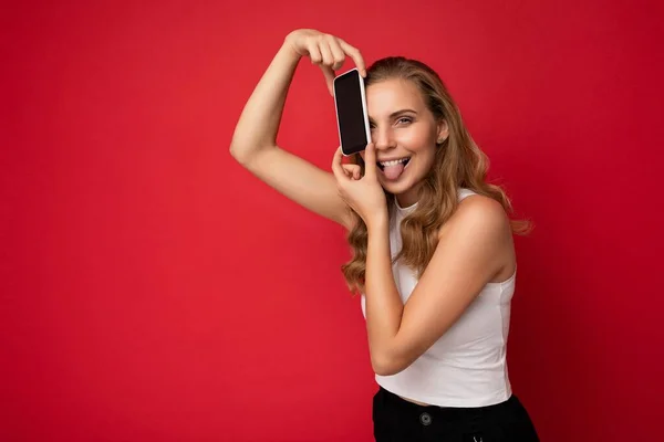 흰색 티셔츠를 입고 빨간 배경에 고립된 금발의 젊은 여성 이 스마트폰을 손에 들고 카메라를 보기 위해 빈 화면을 보여 주는 스마트폰을 들고 있는 사진. — 스톡 사진