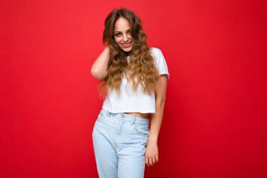 Genç, kıvırcık, seksi ifadeli, neşeli ve mutlu yüzlü beyaz tişörtlü, kırmızı arka planda izole edilmiş, fotokopi alanı olan bir kadın.